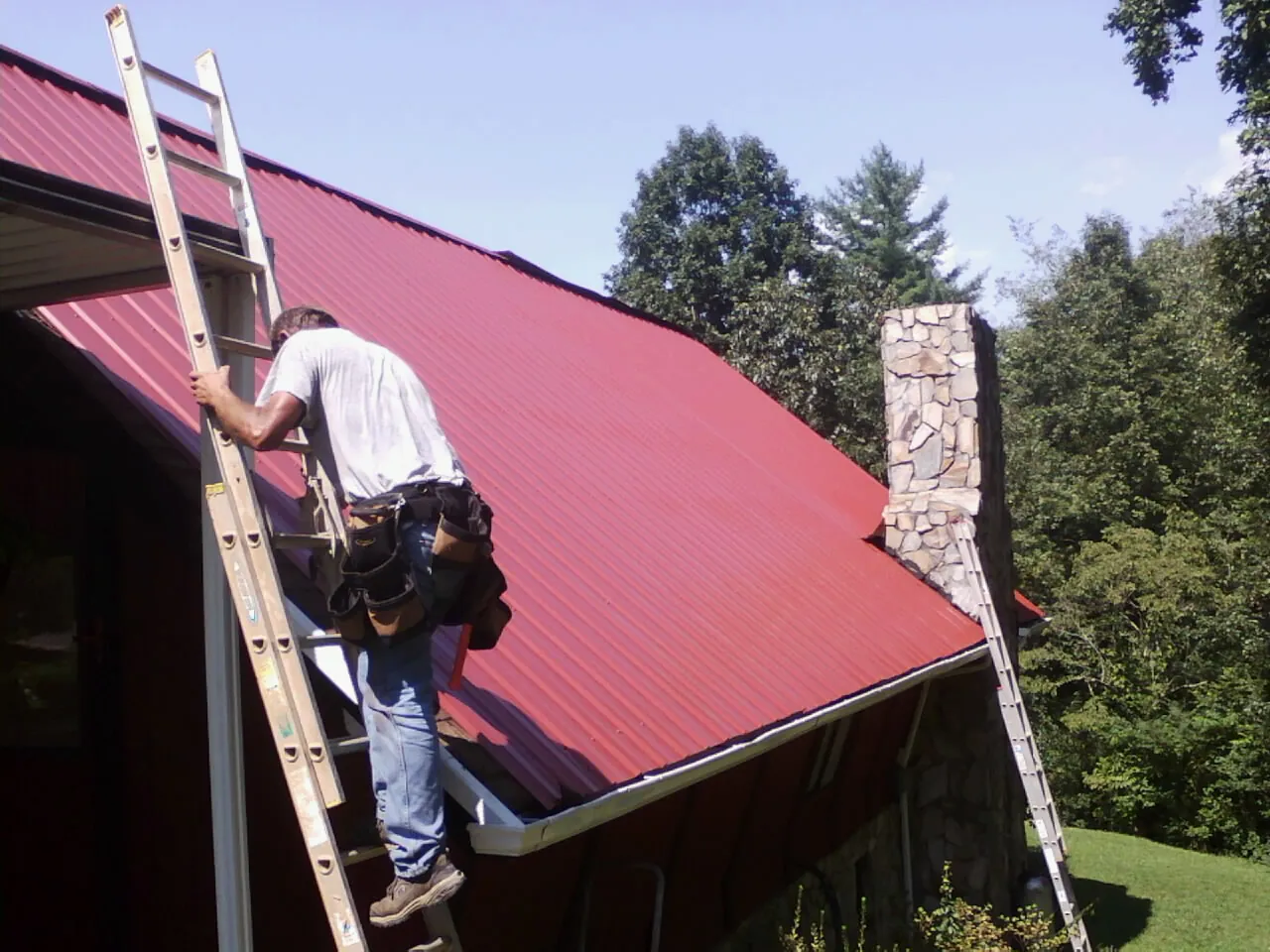 Roofer on a ladder beside a red metal roof and stone chimney — Soffit & Fascia Repair by Roofing Brothers in Round Lake